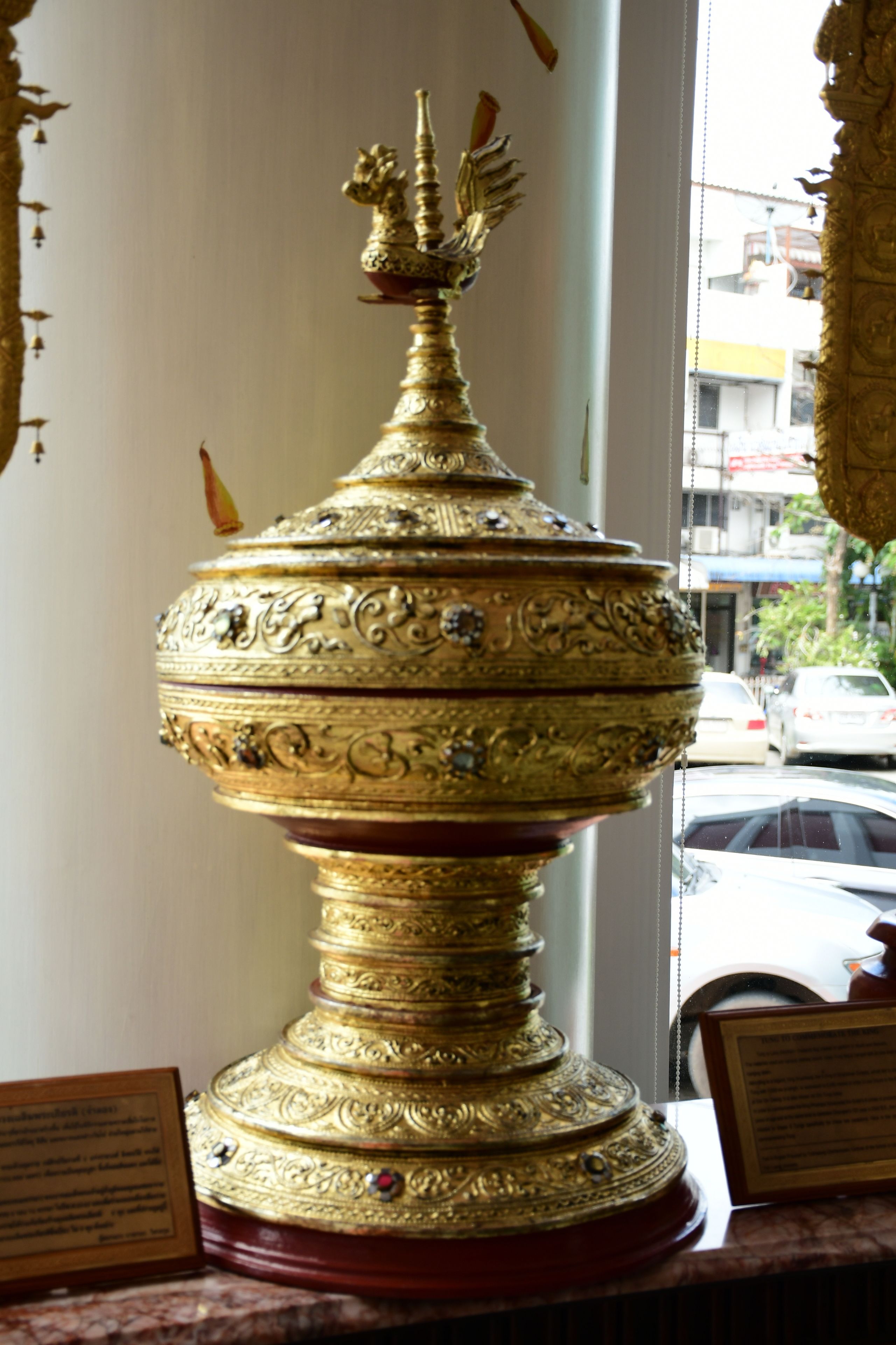 Un objeto dorado ornamentado con diseños intrincados, posiblemente una reliquia religiosa, exhibido en un interior con vistas a una calle.