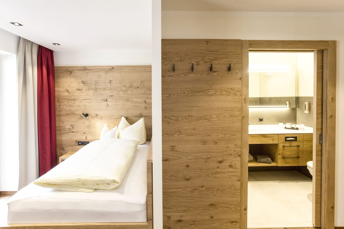 Een hotelkamer met een houten hoofdeinde, een bed met witte lakens, een rood gordijn en een houten garderobe.