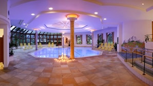 Amplia zona de piscina interior con una columna central, sillas amarillas y una escalera que lleva a un balcón.