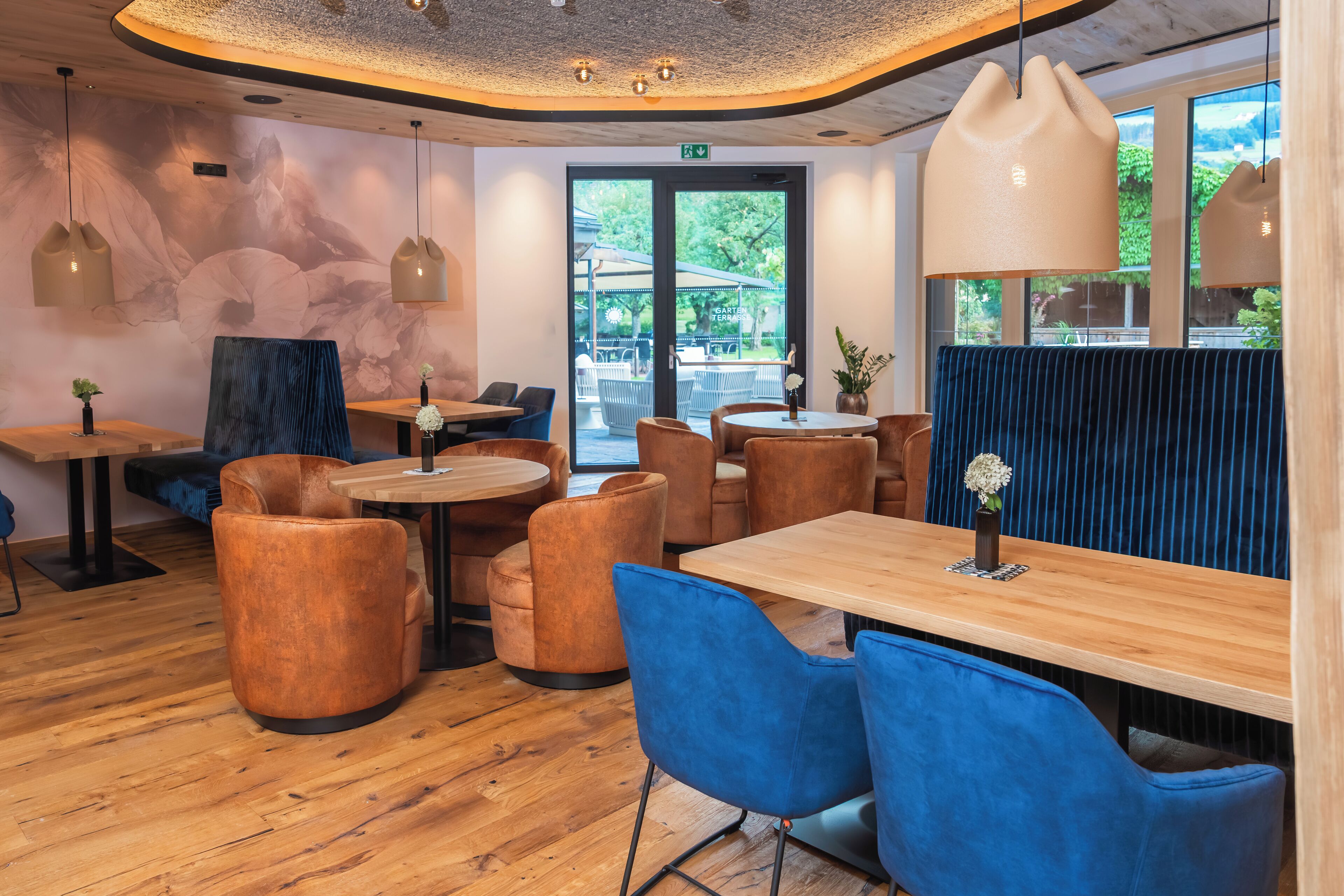 Un interior moderno con suelo de madera, mesas redondas, sillas tapizadas en azul y marrón, e iluminación colgante.