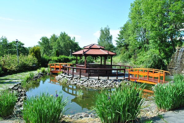 A gazebo with a tiled roof, surrounded by a pond, a wooden bridge, and lush greenery. - Hotel Belweder (Ustron)