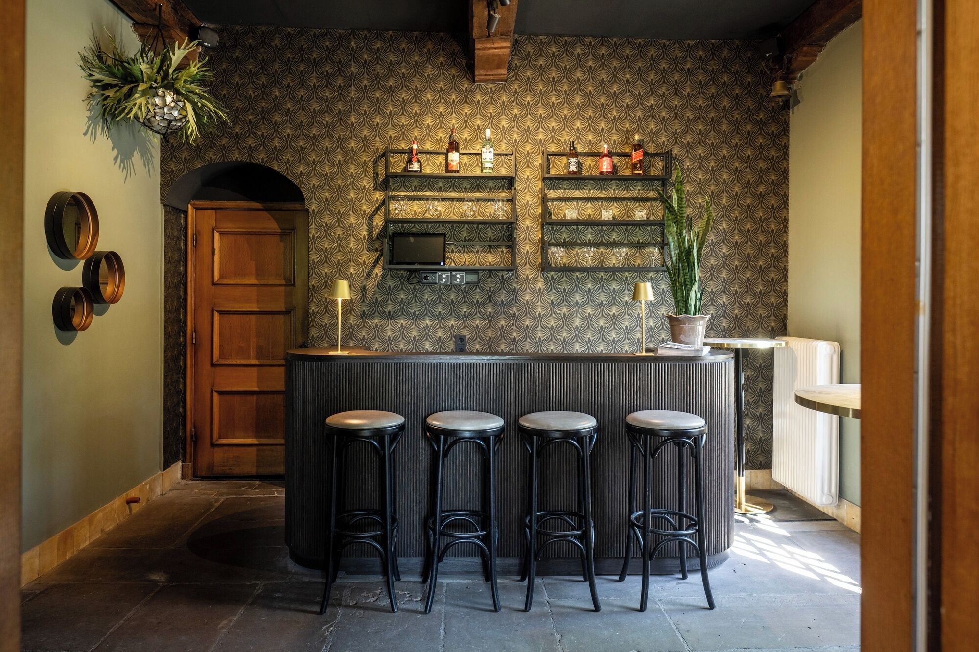 A bar area with a patterned wallpaper, a television, and multiple shelves with bottles. There are four stools in front of the bar.