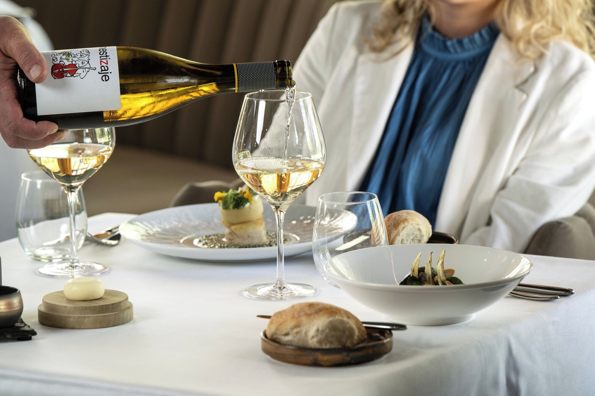 Un tavolo da pranzo con una bottiglia di vino che viene versata in un bicchiere, una ciotola di cibo e un piatto con una piccola ciotolina.