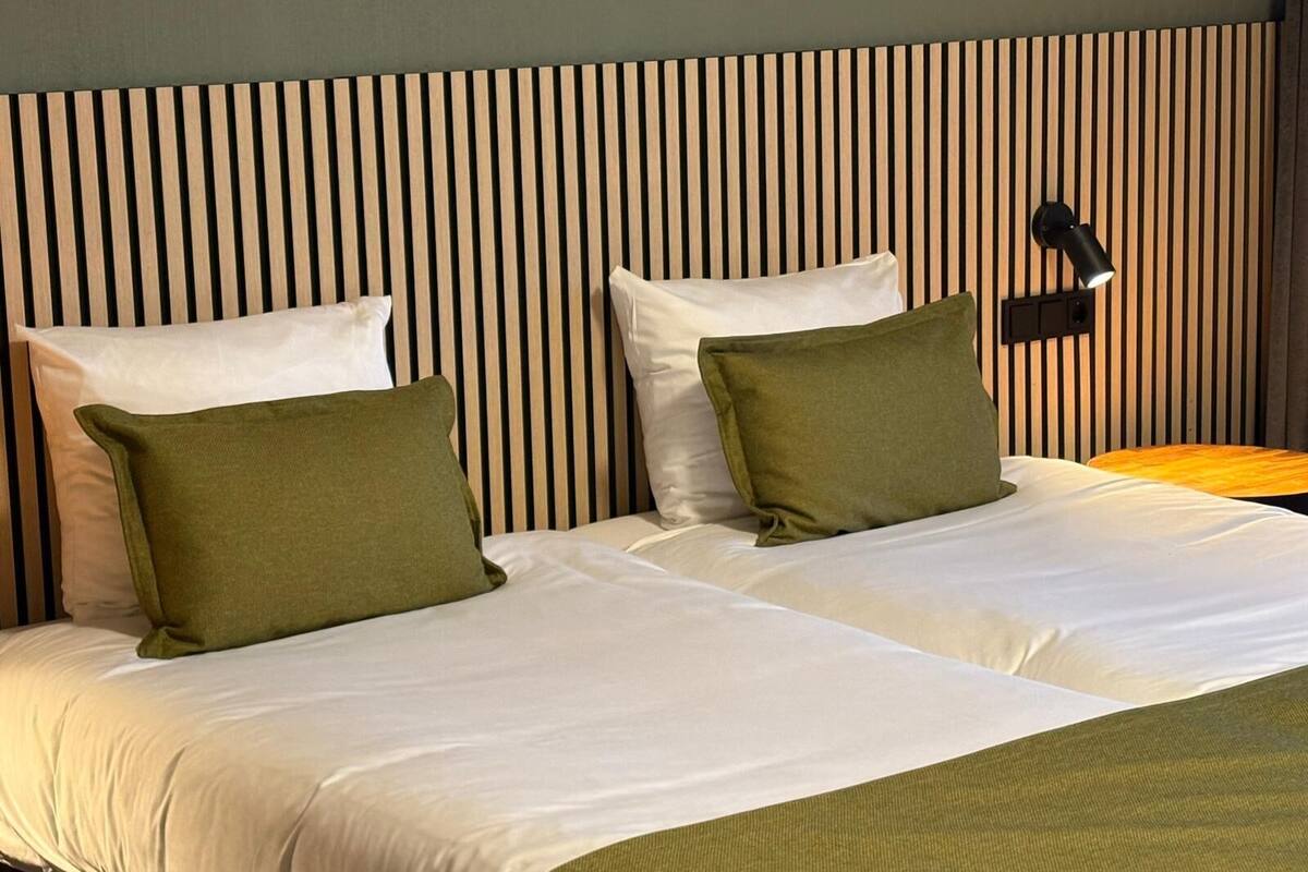 Habitación de hotel con cama, dos almohadas, mesita de noche y lámpara de pared.