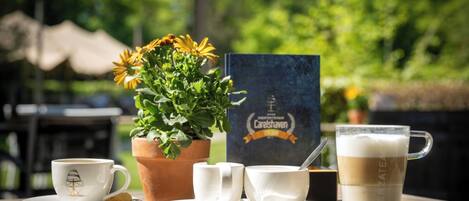 Una mesa con una taza de café, dos platitos, un plato con una galleta, un vaso de leche, un vaso de café, una planta en maceta con flores amarillas y un menú.