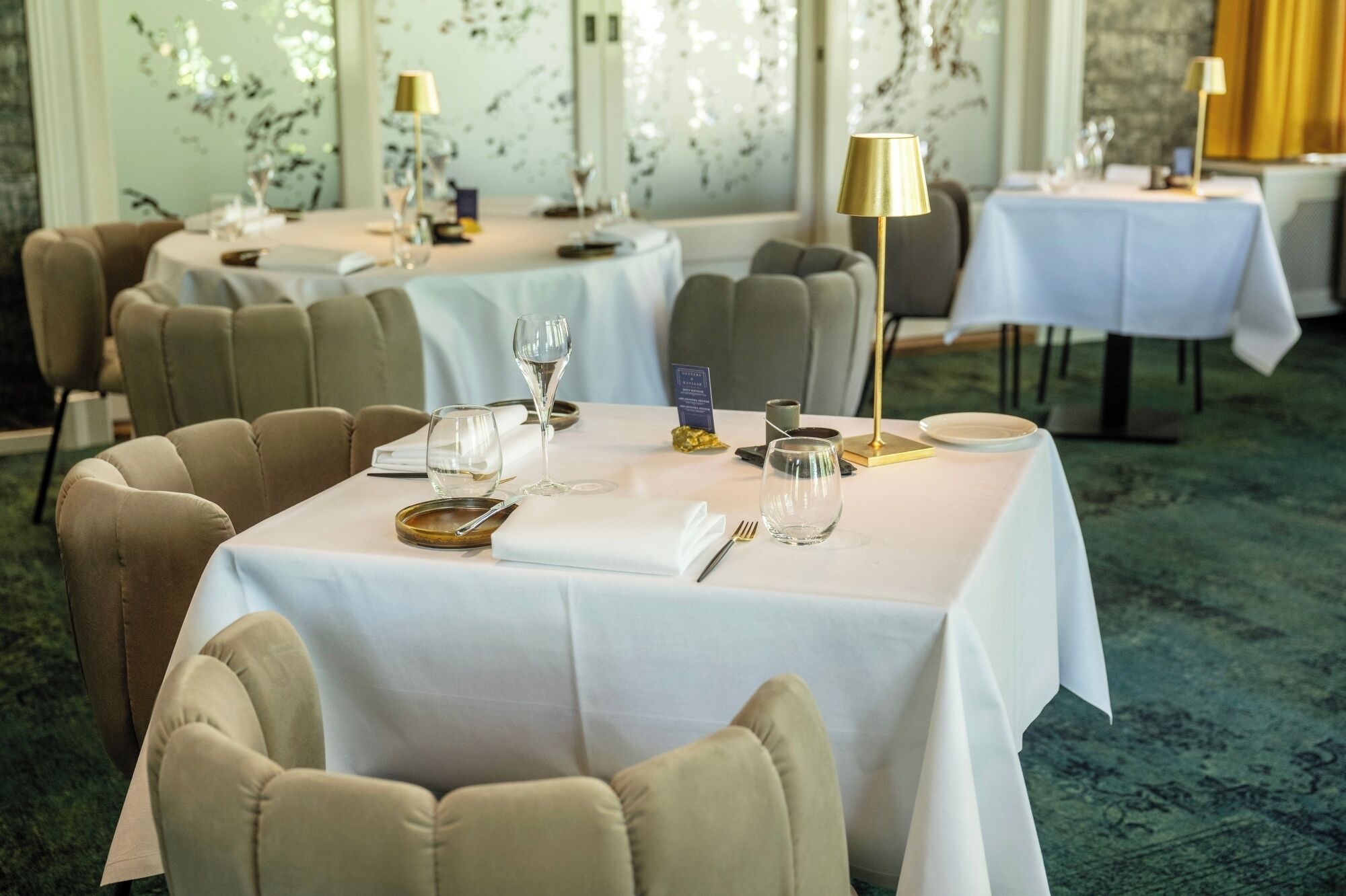 Un restaurante con mesas redondas dispuestas para comer, cada una con mantel blanco, un centro de mesa dorado y una copa de vino.