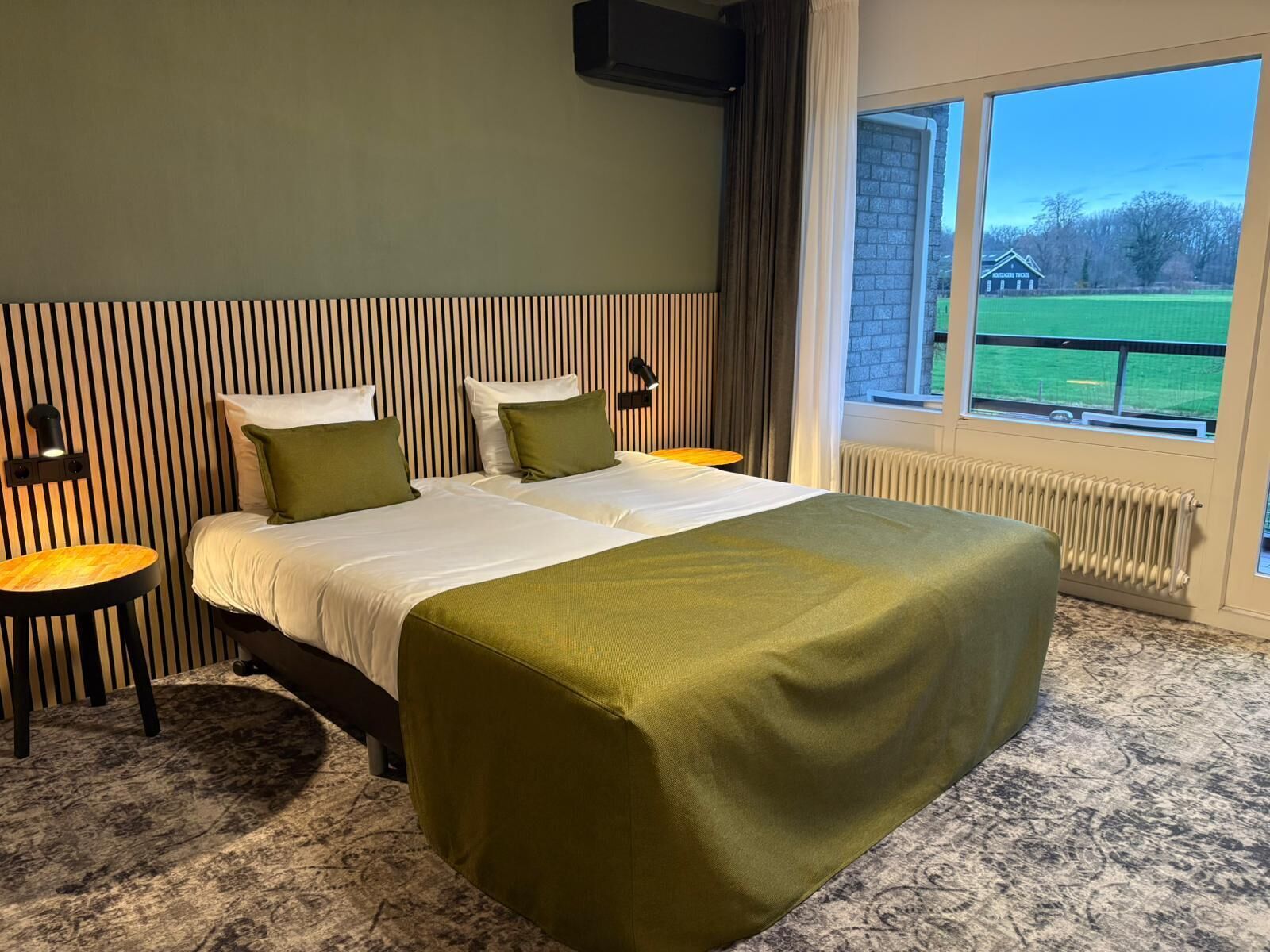 A hotel room with a large bed, two bedside tables, a window with a view of a green field and a building, and a striped wall.