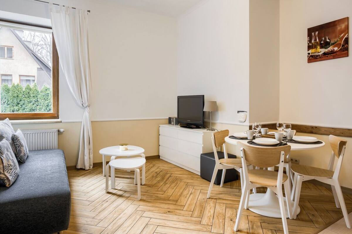 A compact room with a sofa, a small dining table with chairs, a TV on a cabinet, and a window with curtains.