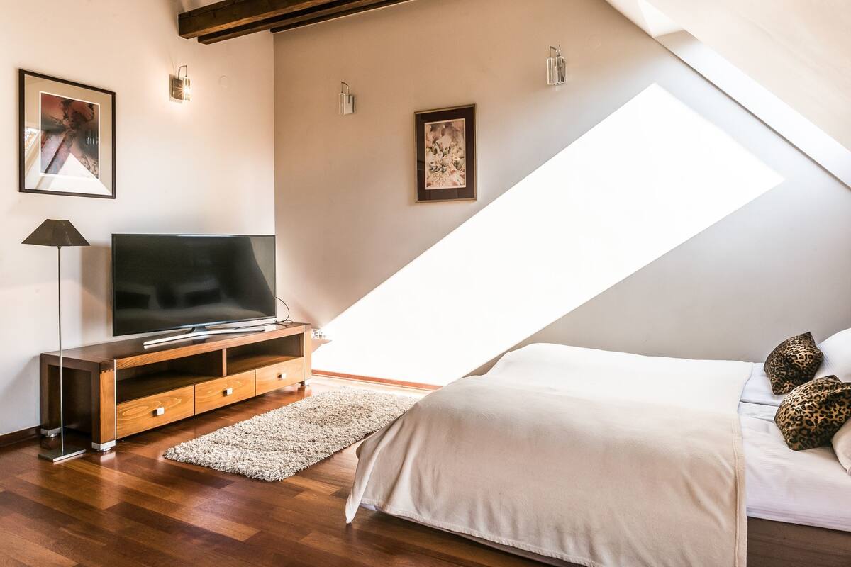 A bedroom with a sloped ceiling, a TV on a wooden stand, a bed with white bedding, and two leopard print pillows.