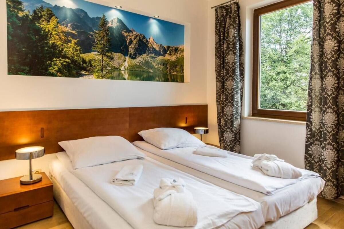 A hotel room with two beds, a large mountain landscape painting, a window with curtains, and a nightstand with a lamp.