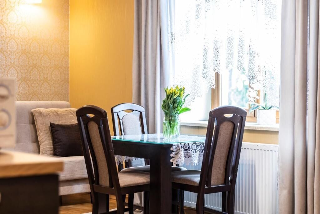 A dining area with a glass table, four chairs, a vase with flowers, a sofa with cushions, and a window with curtains.