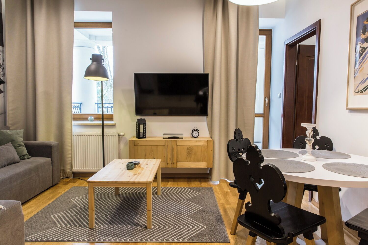 Ein modernes Wohnzimmer mit Fernseher, Holzmöbeln, einem Esstisch mit Stühlen und einem Fenster mit Vorhängen.