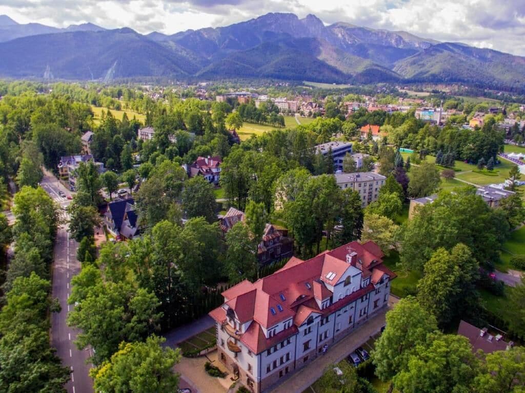 An aerial view of a town with a prominent building featuring a red roof and multiple windows, surrounded by lush greenery and mountains in the background.