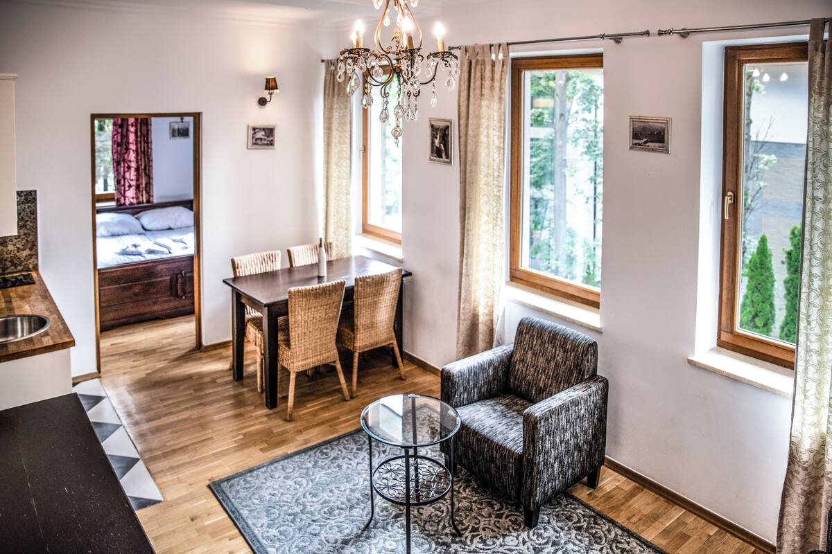 A room with a dining table, chairs, a sofa, a chandelier, and large windows.