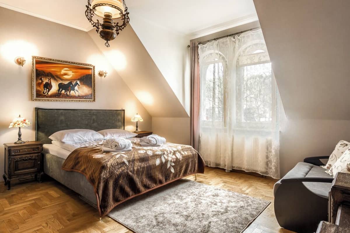 A bedroom with a bed, a nightstand, a lamp, a painting, and a window with curtains.
