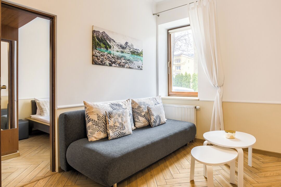A modern room with a grey sofa, a small white table, a painting of a mountain landscape, and a window with curtains.