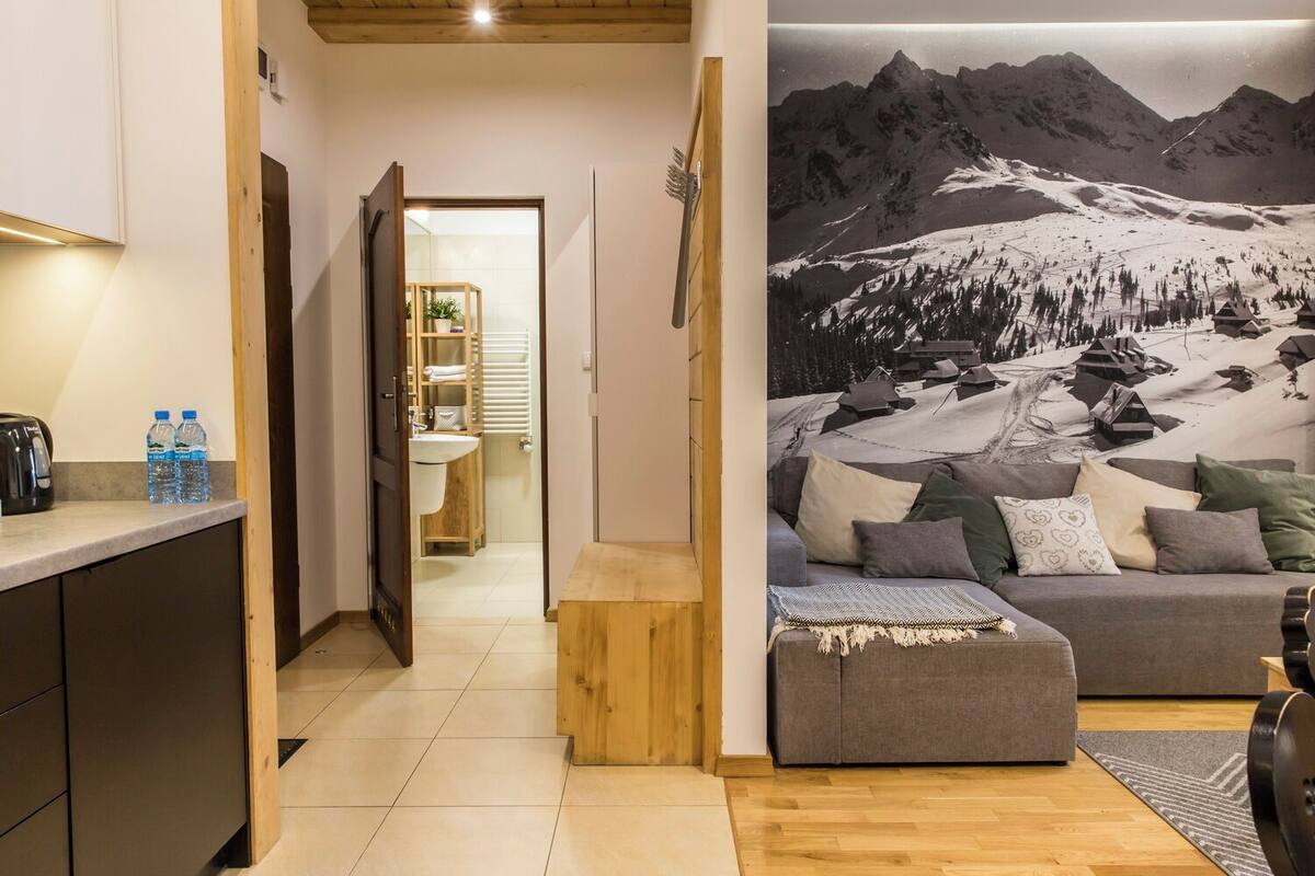 A modern hotel room with a wooden floor, a grey sofa with cushions, a small wooden table, a kitchen area with a kettle and water bottles, and a bathroom with a sink and towel rack.