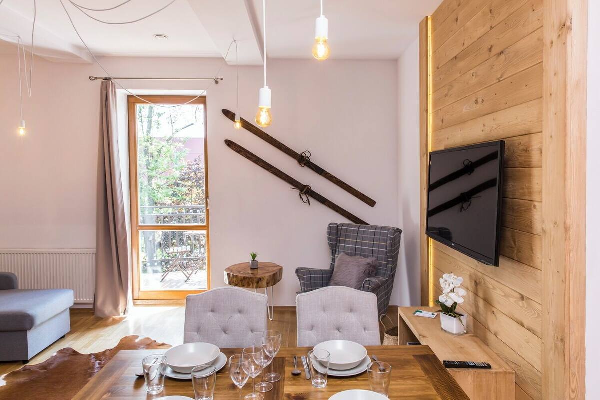 A dining area with a wooden table set for four, a television mounted on a wooden wall, and a window with a view of trees.