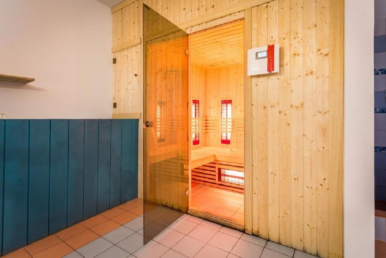 A sauna room with wooden walls and a glass door, a control panel on the wall, and a tiled floor.