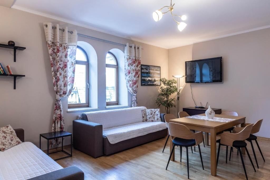 A cozy living room with a sofa, a dining table with chairs, a TV mounted on the wall, and a window with curtains.