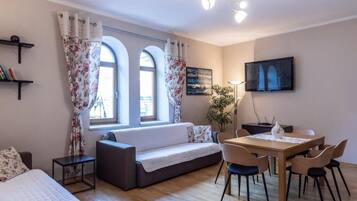 A cozy living room with a sofa, a dining table with chairs, a TV mounted on the wall, and a window with curtains.