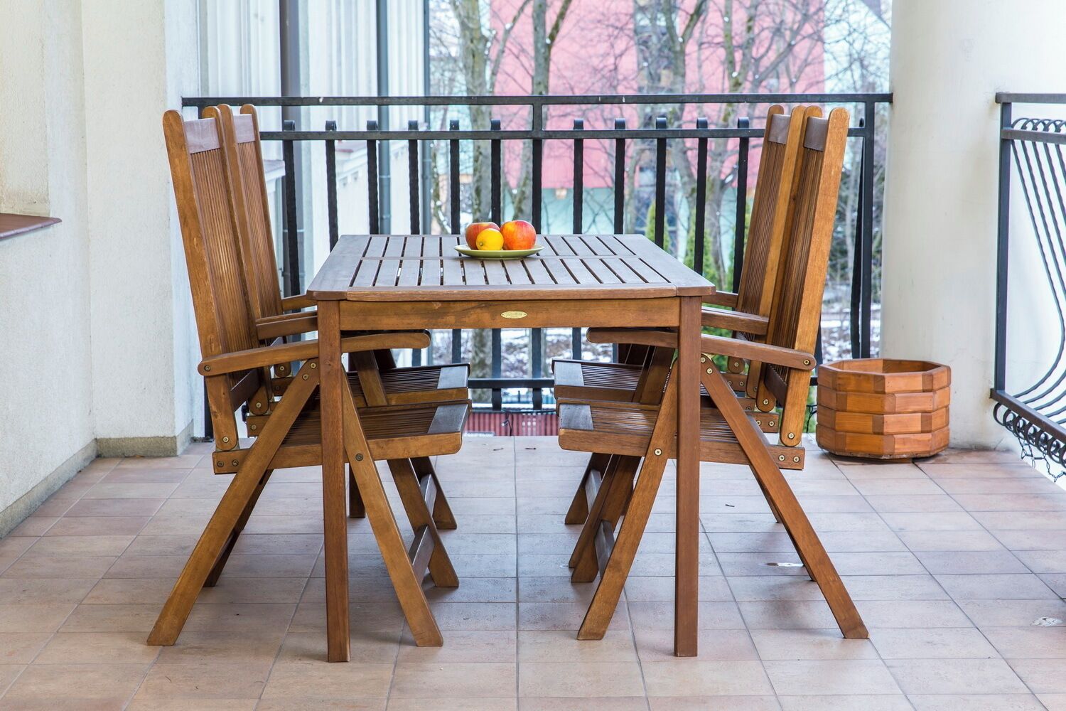 Une table en bois avec quatre chaises, une assiette de fruits et un panier en métal sur un balcon doté d’une balustrade.