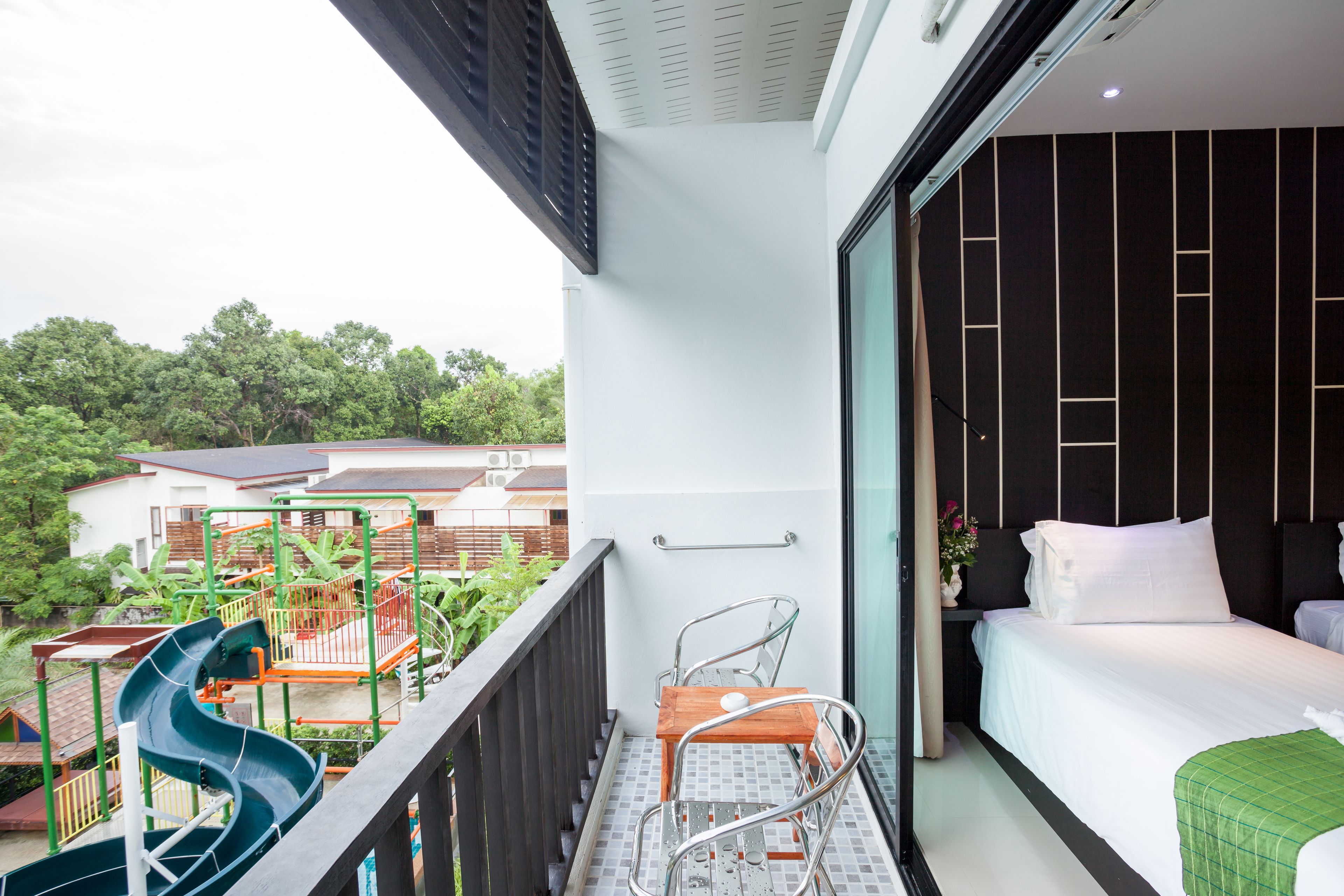 Un balcón con un tobogán, una cama con ropa blanca y vistas a zonas verdes y edificios.