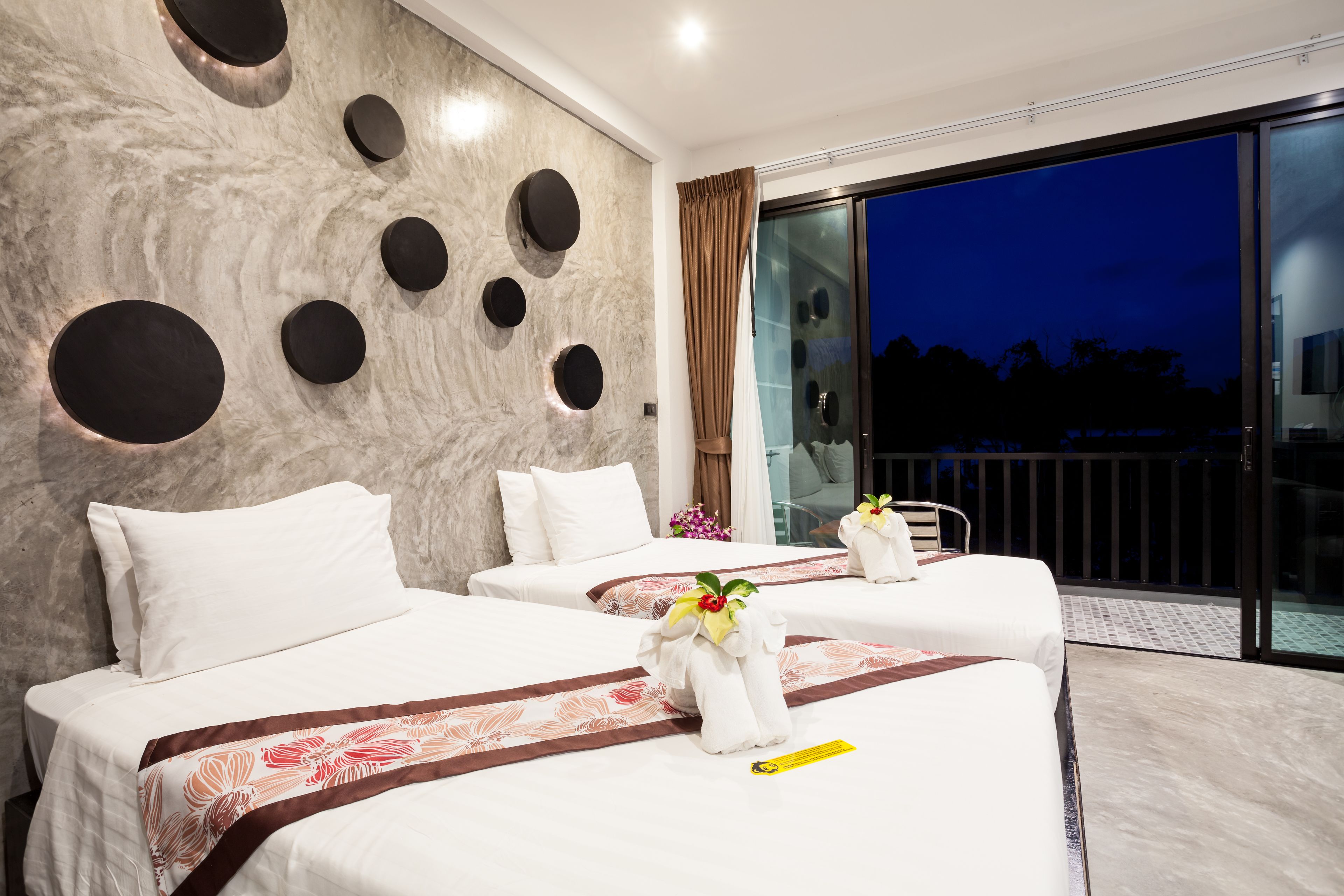 A hotel room with three beds, a marble wall with circular cutouts, a large sliding glass door to a balcony, and a flower arrangement on the beds.