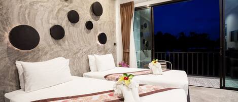 A hotel room with three beds, a marble wall with circular cutouts, a large sliding glass door to a balcony, and a flower arrangement on the beds.