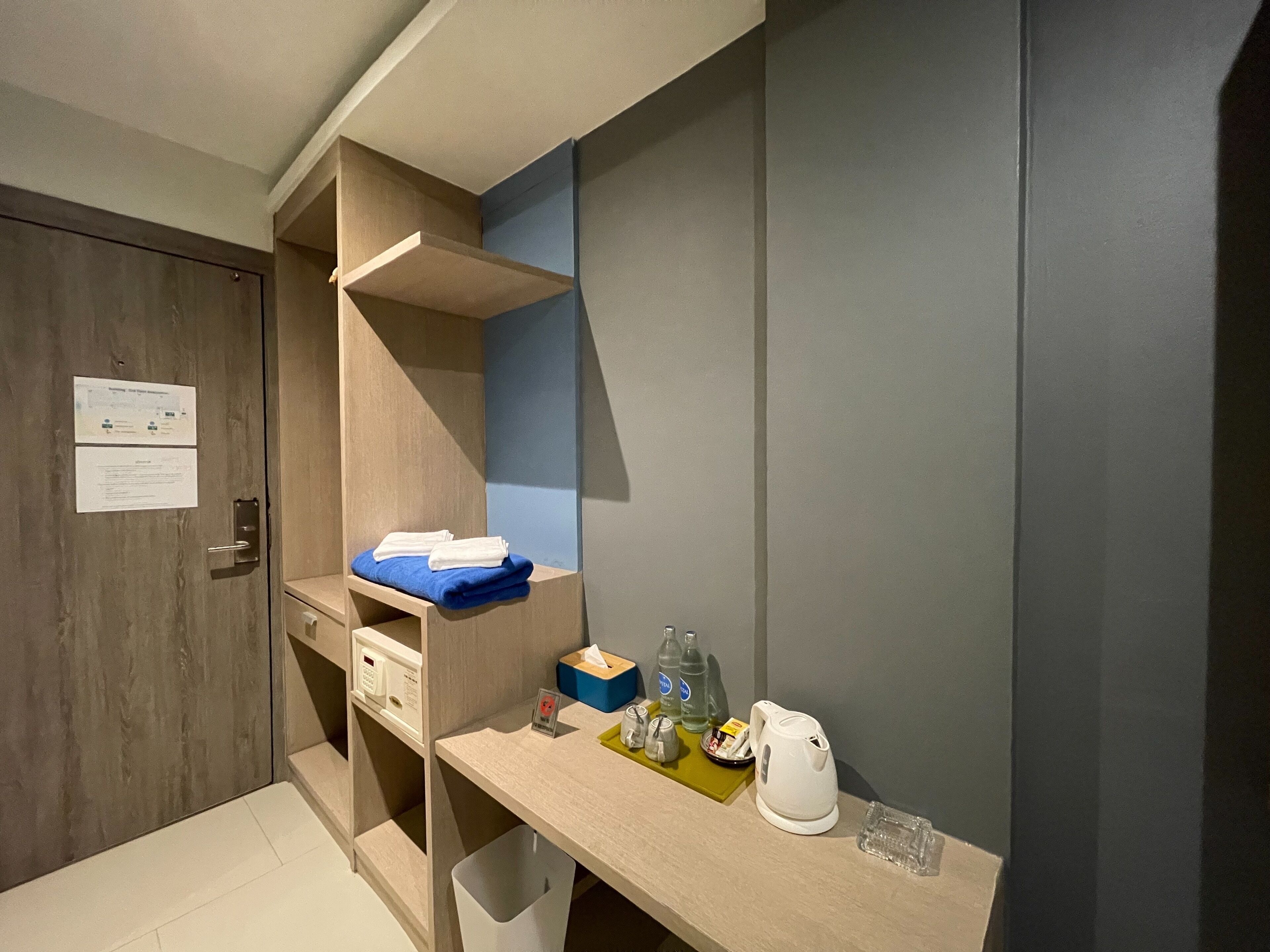 A compact hotel room with a wooden door, a small desk with a kettle, bottles, and a tissue box.