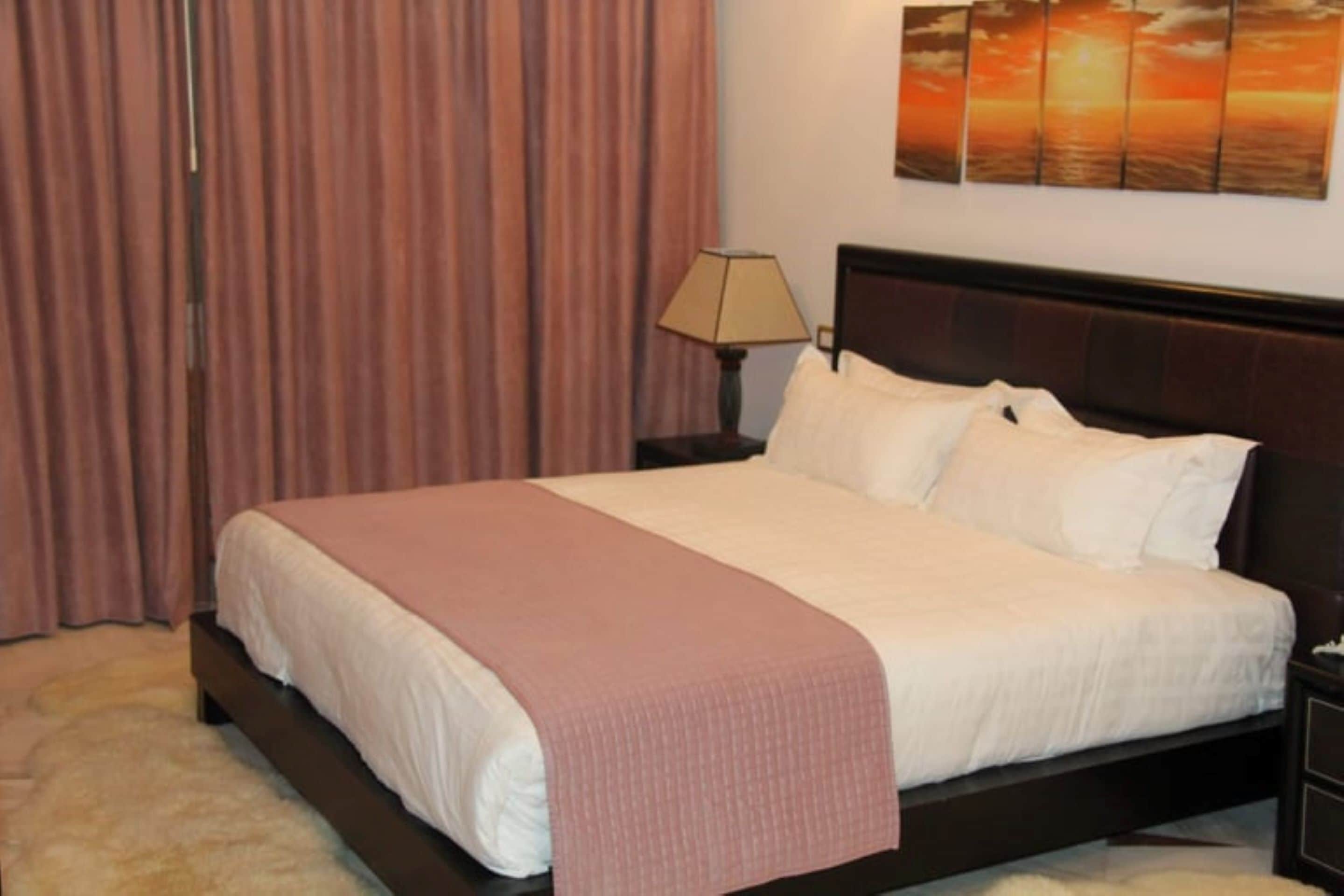 A neatly made bed with a pink blanket, a bedside lamp, and a sunset painting on the wall.