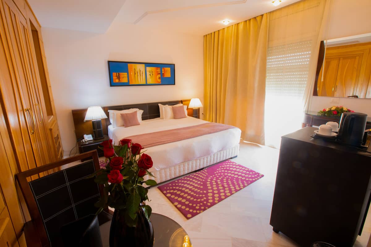 A hotel room with a bed, bedside lamps, a desk, a vase with flowers, and a large window with curtains.