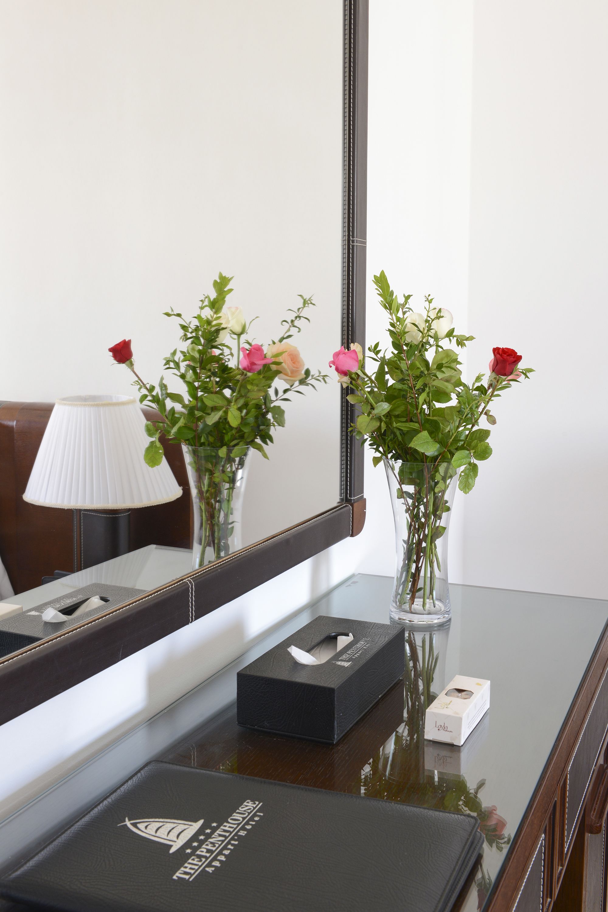 Ein Hotelzimmer mit Glastisch, Blumen in einer Vase, einer Lampe und einem Spiegel, der das Zimmer reflektiert.
