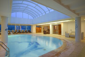 Una amplia piscina cubierta con techo de cristal, sillas de descanso y una zona de recepción.