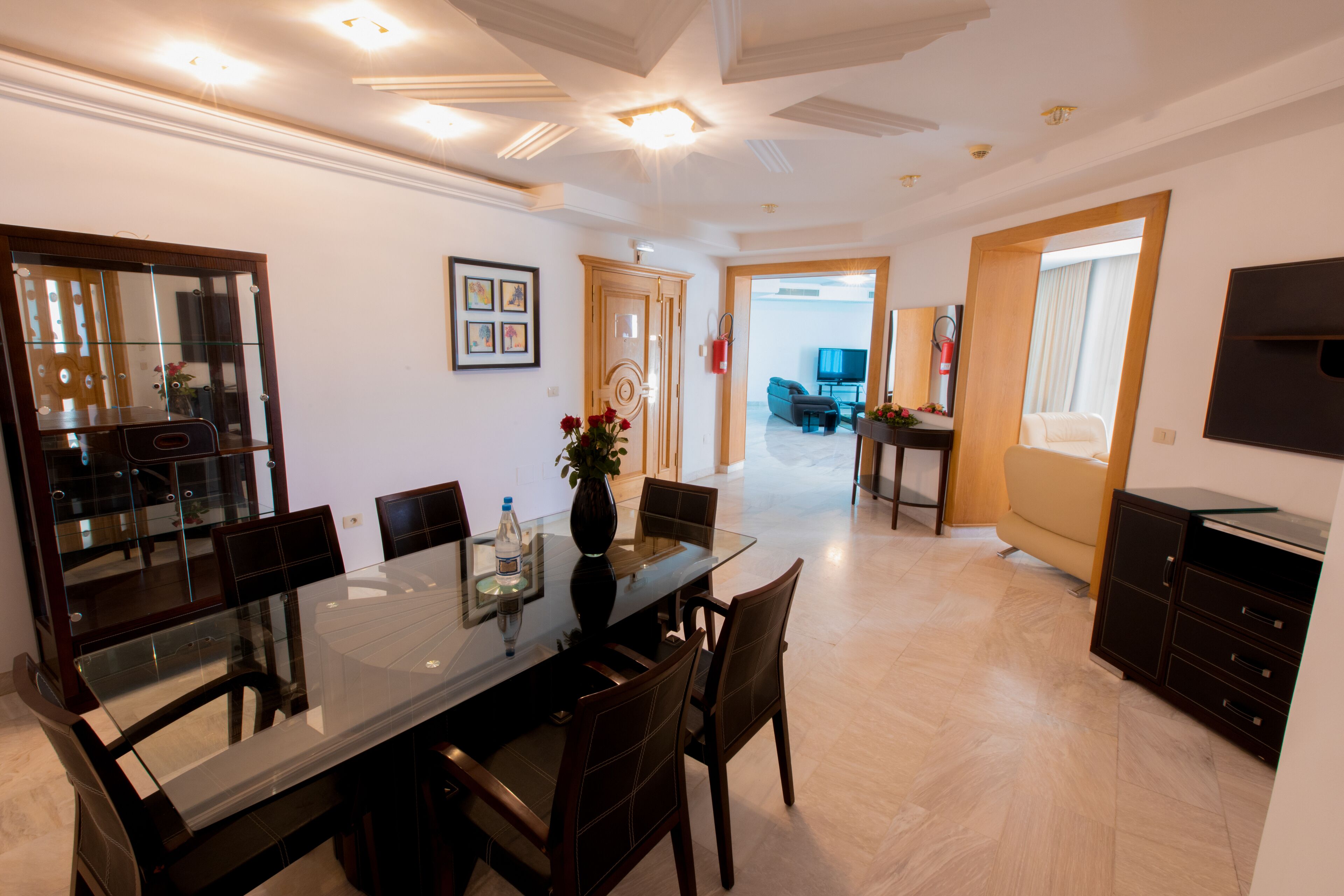 A modern dining area with a glass table and wooden chairs, a vase with flowers, and a large mirror on the wall.