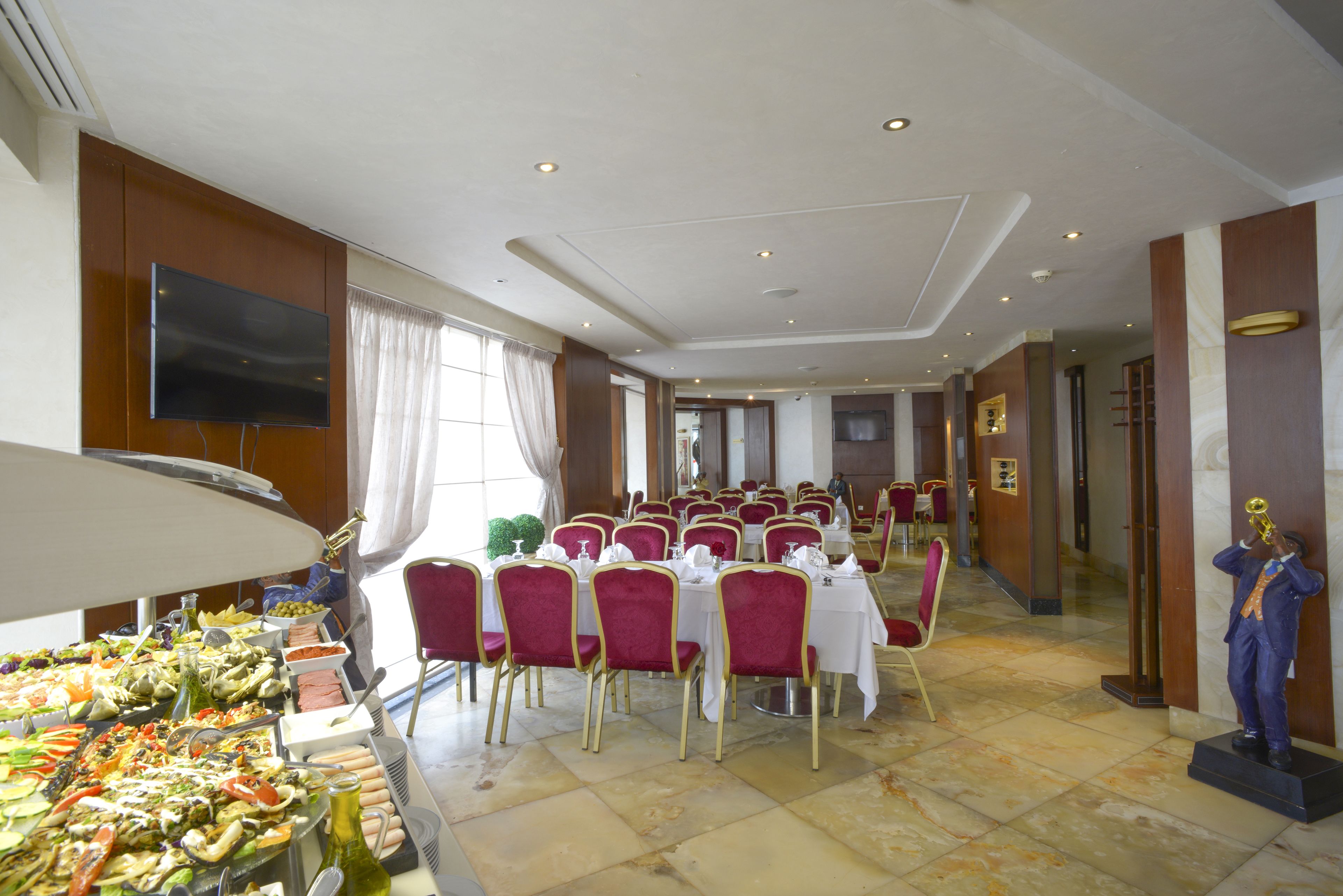 Une salle de banquet avec des rangées de tables et de chaises, un espace buffet proposant divers plats, et une statue représentant une personne jouant de la trompette.