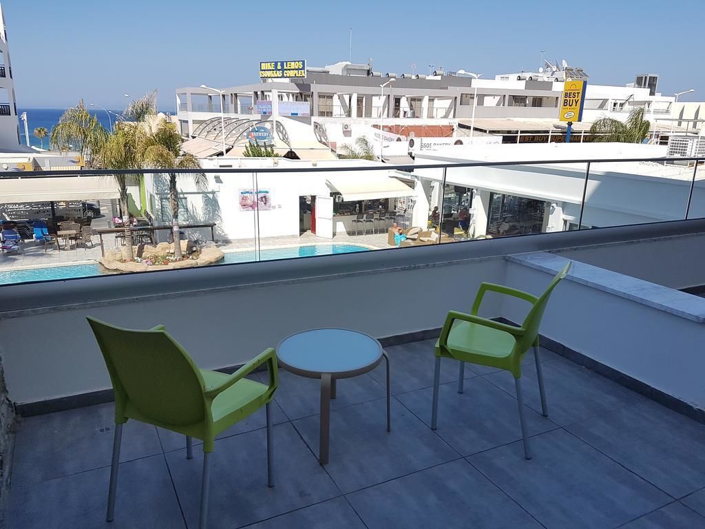 A rooftop terrace with two green chairs and a small table, overlooking a pool and buildings with signage.