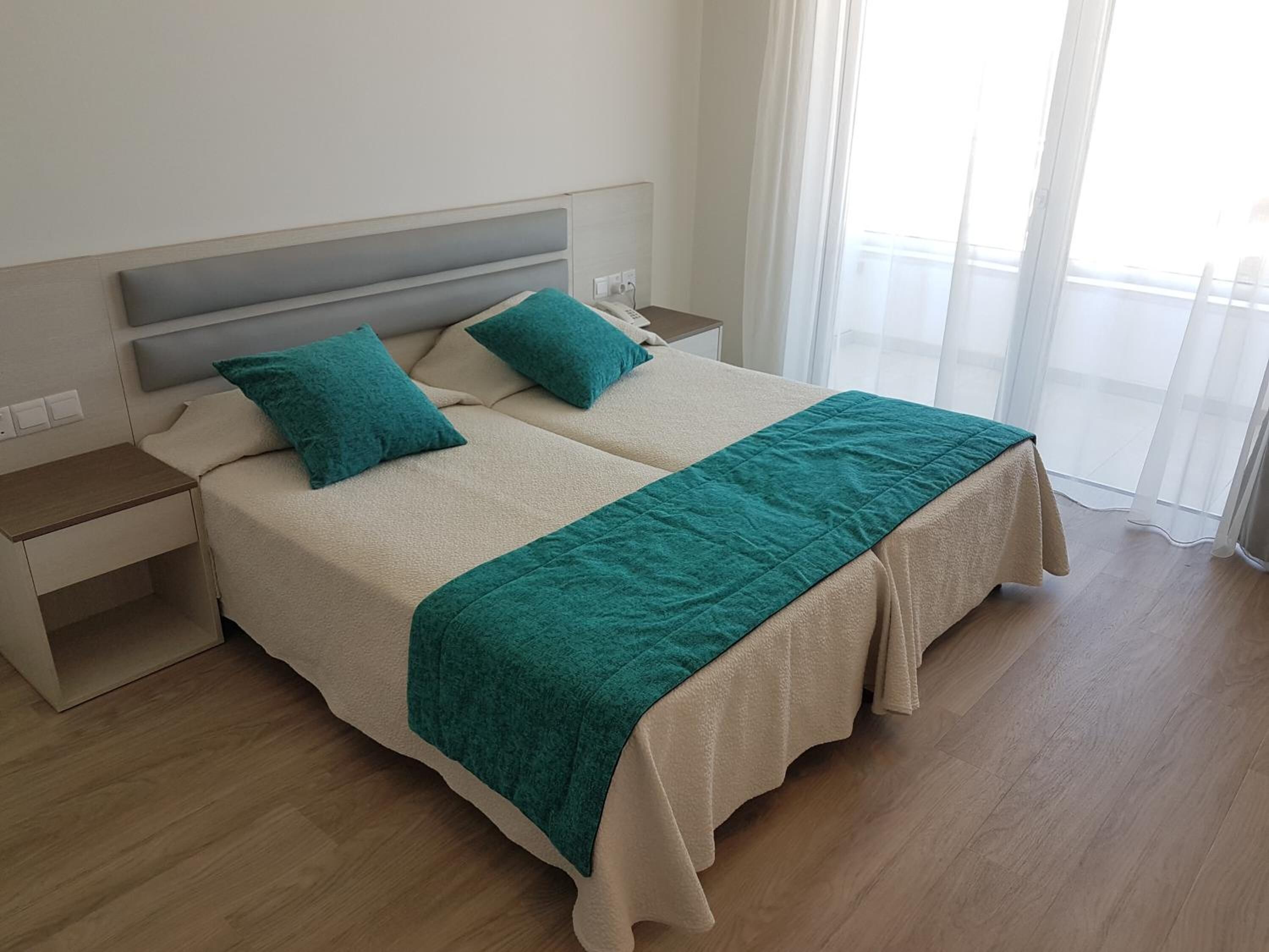 A double bed with a beige headboard and two pillows, a green throw blanket, and a nightstand with a lamp.