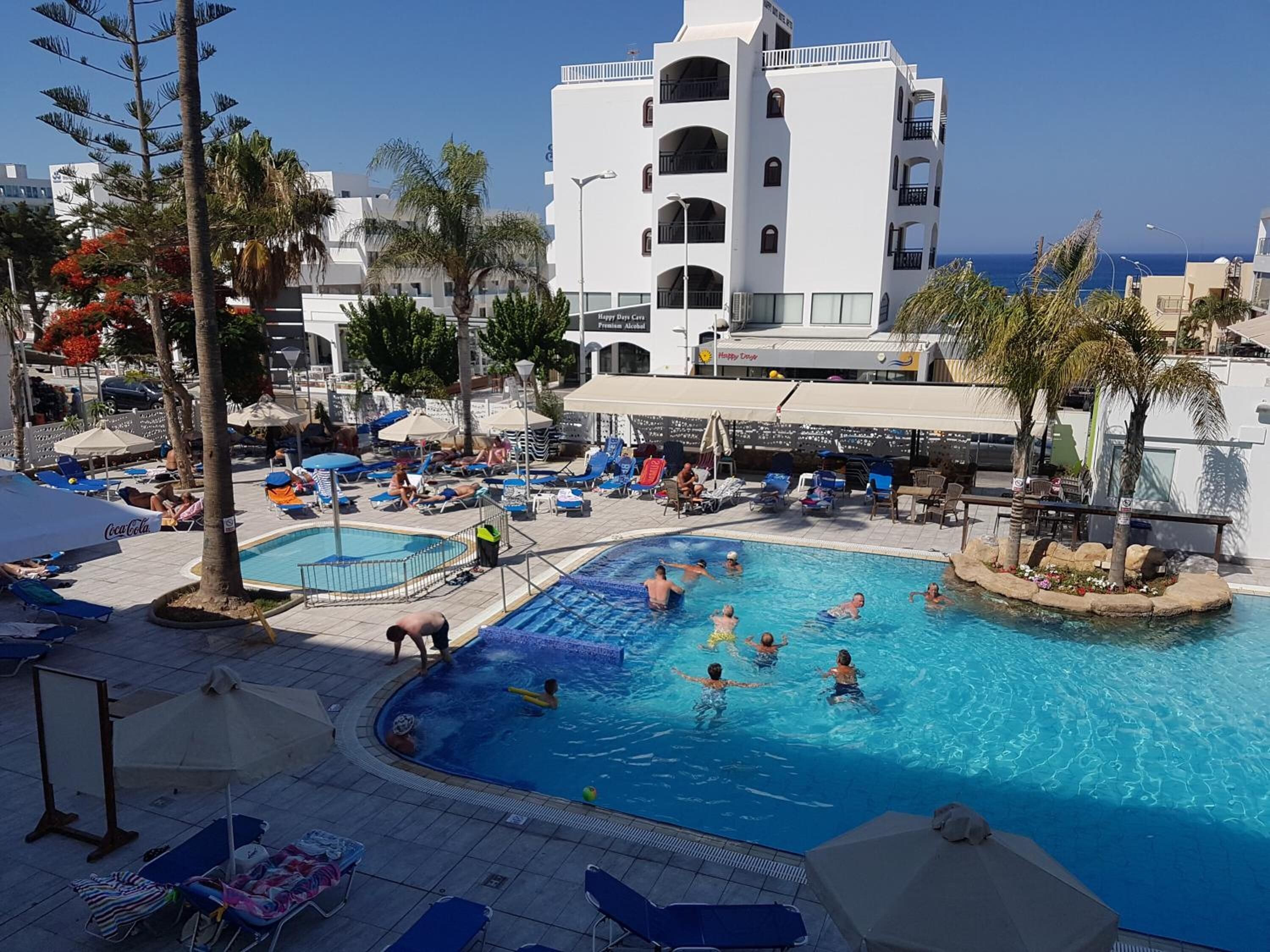 A swimming pool area with lounge chairs and umbrellas, surrounded by palm trees and a white building with balconies.