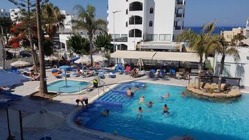 A swimming pool area with lounge chairs and umbrellas, surrounded by palm trees and a white building with balconies.