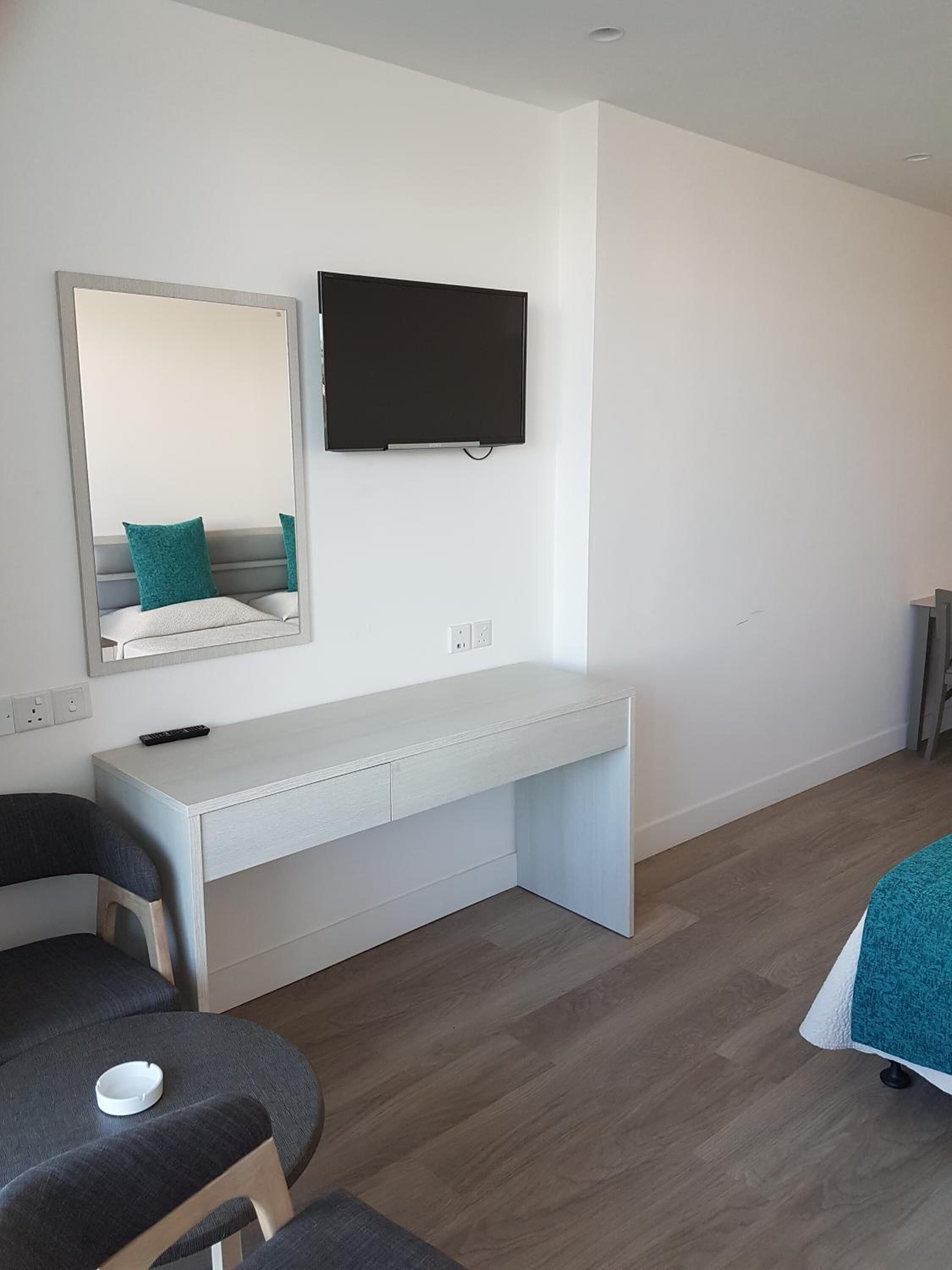 A modern room with a white desk, a television mounted on the wall, a mirror above the desk, and a grey sofa with a remote control on it.