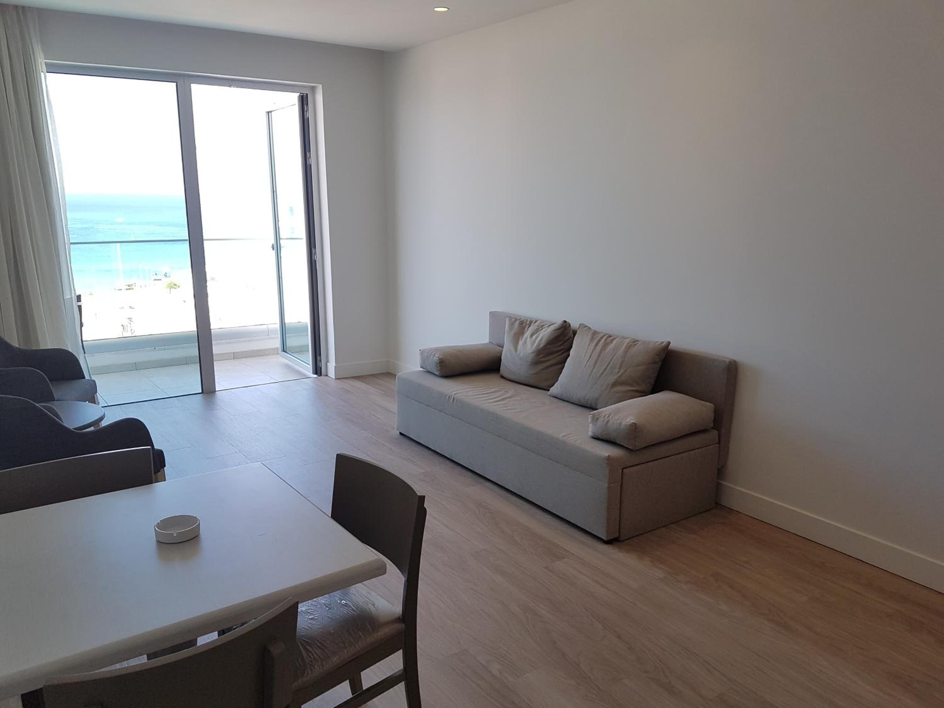 A modern living room with a sofa, a dining table, and a chair. There is a large window with a view of the ocean.