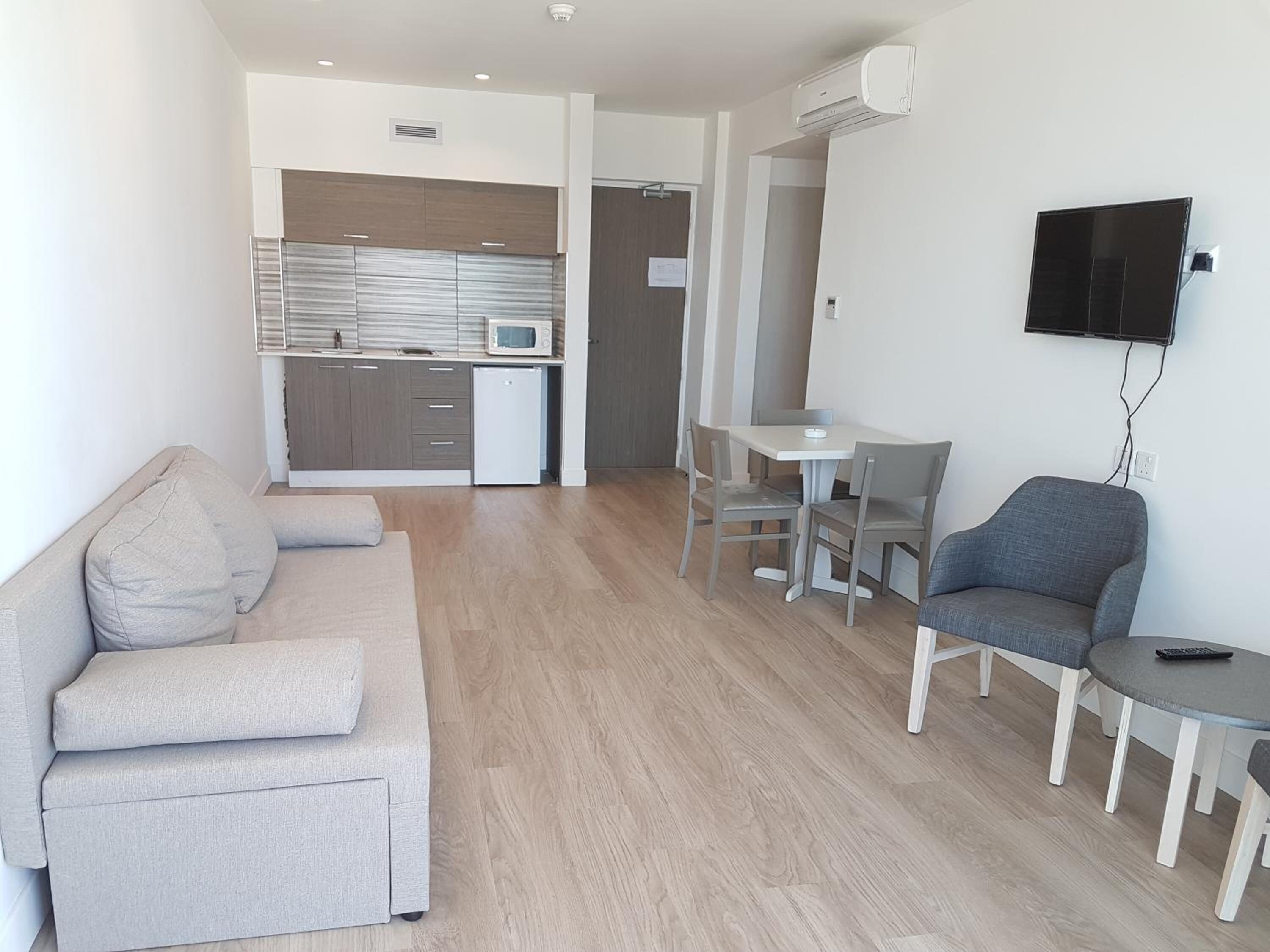 A modern living room with a sofa, a dining table with chairs, a TV mounted on the wall, and a kitchen area in the background.