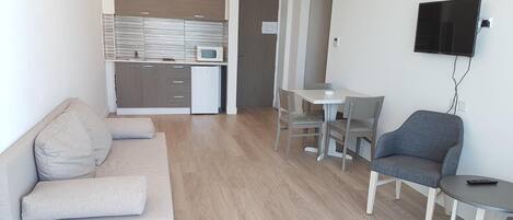 A modern living room with a sofa, a dining table with chairs, a TV mounted on the wall, and a kitchen area in the background.