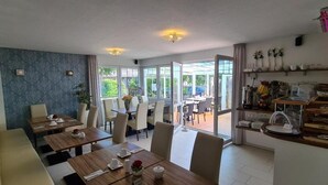 Ein Essbereich mit Tischen und Stühlen, eine Küchenzeile mit Arbeitsfläche und Geräten sowie ein Blick ins Freie durch große Fenster.