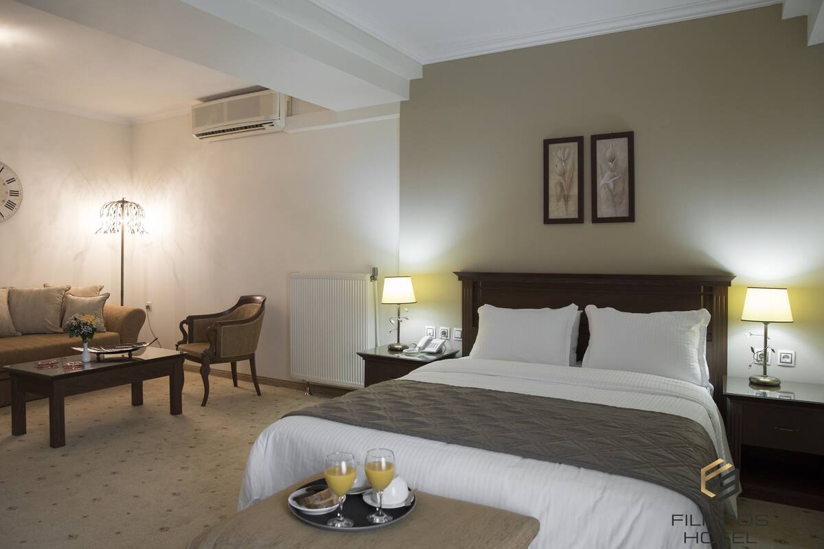 A hotel room with a large bed, a sitting area with a sofa and armchair, a coffee table, and a tray with glasses of juice.