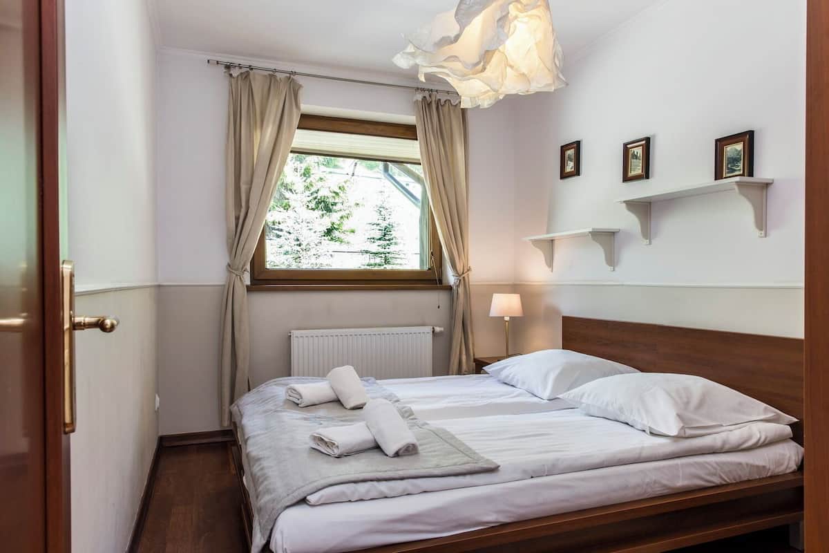 Una cama bien hecha con sábanas blancas, una ventana con cortinas, una lámpara de noche y un estante con fotografías.