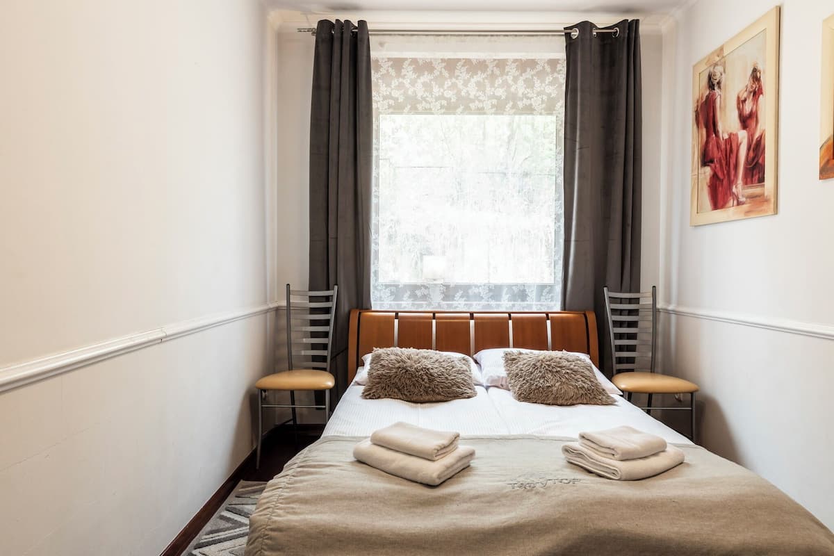 Una habitación de hotel con una cama, dos sillas, un ventanal con cortinas y un cuadro en la pared.