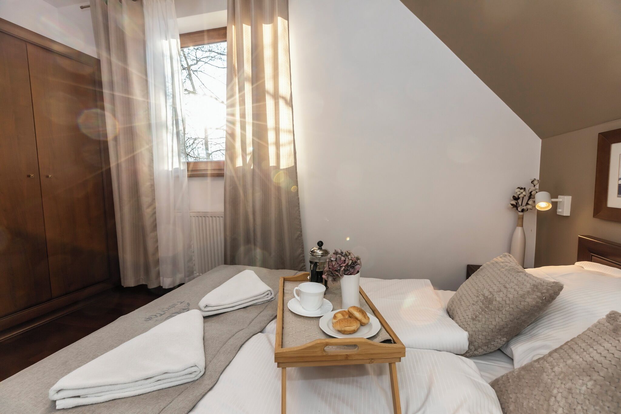 Hotellihuone, jossa on sänky, puinen vaatekaappi, verhoilla varustettu ikkuna, yöpöytä maljakolla ja tarjotinalusta, jossa on kuppi ja leivonnaisia.
