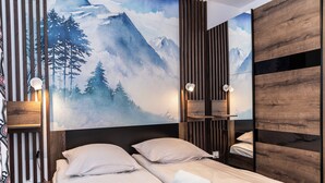 Habitación de hotel con cama, mesita de noche, lámpara de pared y un cuadro grande de un paisaje montañoso.